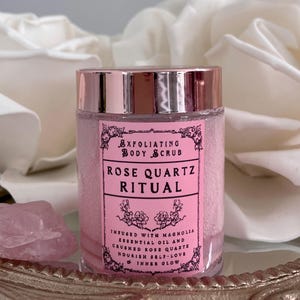 May include: A pink exfoliating body scrub jar with a rose gold lid and label that reads "Rose Quartz Ritual." The jar is filled with pink granules and sits on a decorative tray. A rose quartz crystal and white roses are in the background.