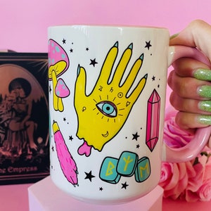 May include: White ceramic mug with a pink handle, featuring a yellow hand with an eye, mushrooms, a pink crystal, and runes. The mug is decorated with stars and other mystical symbols. The background is pink.