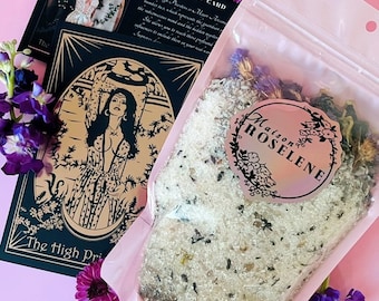 May include: A pink-tinted package of bath salts with the text "Maison Roselene" on a sticker, next to a tarot card with the words "The High Priestess." The card features a gold illustration of a woman and text about the card's meaning. Purple flowers are scattered around.