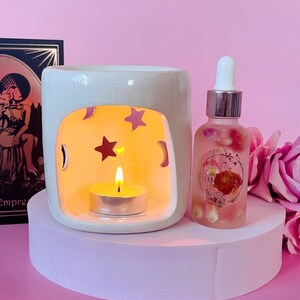 May include: A white ceramic essential oil diffuser with star and crescent moon cutouts, lit by a small candle. A glass bottle of oil with rose petals sits next to it. The items are on a pink platform, with a pink background.
