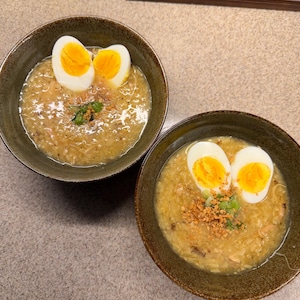May include: Two bowls of congee, a rice porridge, each topped with a halved hard-boiled egg. Garnishes include chopped spring onions and fried garlic. The bowls are a dark brown colour and sit on a light grey surface.