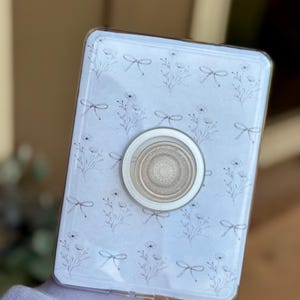 Peut inclure: Coque de téléphone transparente avec un motif floral et libellules. L'étui est doté d'une poignée pop-up argentée au centre. Le motif présente des dessins au trait de fleurs et de libellules sur fond bleu clair.