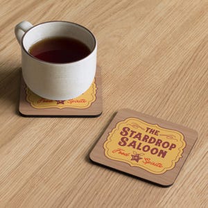 Peut inclure: Deux dessous de verre en bois avec un motif de grain de bois brun clair. L'un des dessous de verre a une tasse blanche avec du th&eacute; dessus. L'autre dessous de verre porte le texte "The Stardrop Saloon Food Spirits" dans un design jaune et rouge.