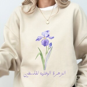 Könnte beinhalten: Ein beiger Pullover mit einem lila Iris-Blumenmotiv und dem Text "الزهور اللوطنية لملابسنا" in arabischer Schrift.
