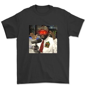 May include: A black t-shirt with a photo of a man wearing a patterned hat and a white shirt. The man has red eyes and is speaking into a microphone. The text "HAG" is visible in the background.