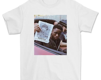 May include: A white t-shirt with a photo of a young man holding a wanted poster. The poster says "WANTED UP TO $5,000 REWARD".