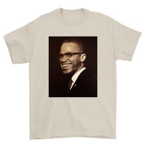 May include: A beige t-shirt featuring a sepia-toned portrait of a smiling man wearing glasses and a suit. The image is centered on the shirt, showcasing the man's face and upper body. The shirt's color provides a neutral backdrop for the portrait.
