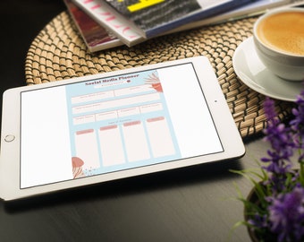 May include: A white tablet with a blue and pink social media planner on the screen. The tablet is on a woven placemat with a cup of coffee and a potted plant with purple flowers.