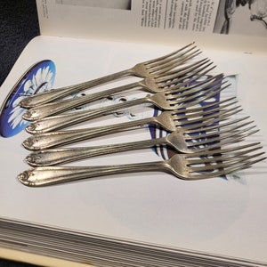 May include: A set of six silver forks with ornate handles. The forks are arranged in a stack, with the handles facing up. The forks are shiny and reflect the light.