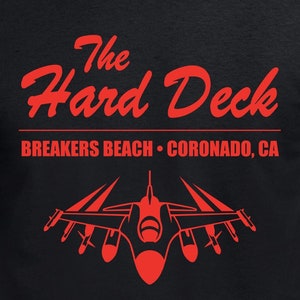 Op de afbeelding: Zwart T-shirt met rode tekst die "The Hard Deck" en "Breakers Beach - Coronado, CA" luidt. Onder de tekst bevindt zich een rood silhouet van een straaljager.