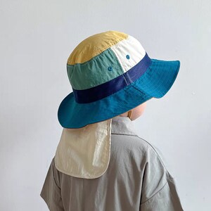 Könnte beinhalten: Ein bunter, mehrfarbiger Sonnenhut im Patchwork-Stil mit einem Nackenschutz. Der Hut ist aus leichtem Stoff und hat eine breite Krempe. Der Hut ist blau, grün, gelb und weiß.