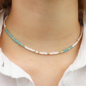 Könnte beinhalten: Eine zarte Choker-Kette mit weißen und türkisfarbenen Perlen. Die Kette ist aus kleinen, gleichmäßig verteilten Perlen gefertigt und hat ein schlichtes, minimalistisches Design.