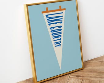 May include: A framed wall art print featuring a cream-coloured triangular pennant on a light blue background. The pennant displays the text LAKE COUNTRY in bold blue capital letters, with an orange horizontal bar at the top of the pennant.