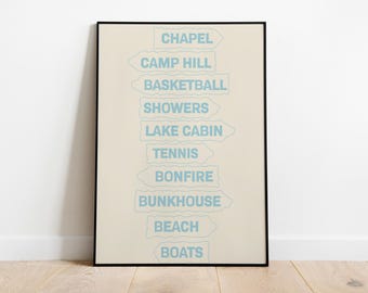 May include: A framed vertical poster with a cream background featuring a stack of light blue directional sign shapes. The text reads: CHAPEL, CAMP HILL, BASKETBALL, SHOWERS, LAKE CABIN, TENNIS, BONFIRE, BUNKHOUSE, BEACH, and BOATS in a sans-serif font.