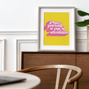 May include: A framed wall art print featuring a bright yellow background with a pink brushstroke graphic and the white text "Pour yourself a cup of Ambition" in a retro-inspired script font, displayed in a light-colored wooden frame.