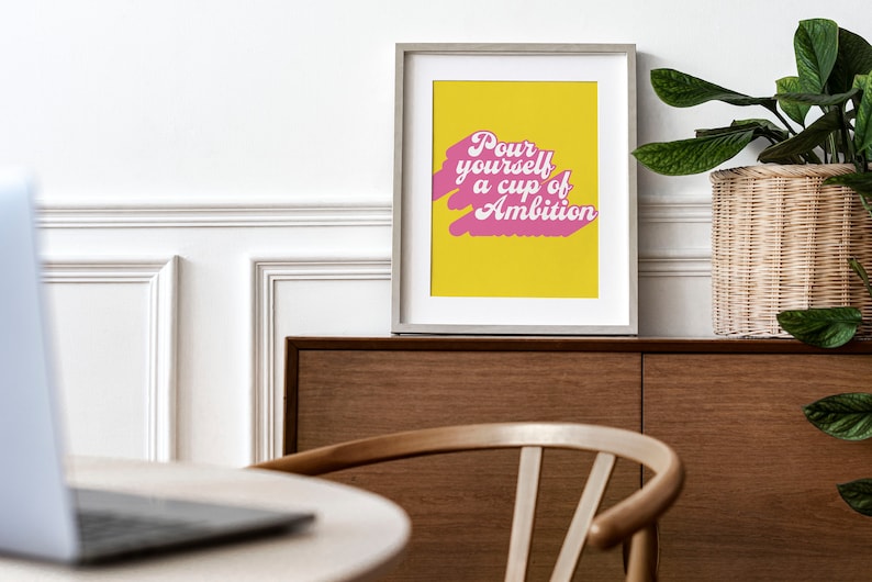 May include: A framed wall art print featuring a bright yellow background with a pink brushstroke graphic and the white text "Pour yourself a cup of Ambition" in a retro-inspired script font, displayed in a light-colored wooden frame.