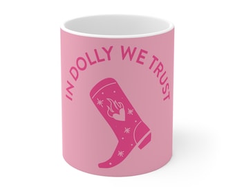 May include: A pink ceramic mug featuring the text IN DOLLY WE TRUST in a curved font above a graphic of a cowboy boot. The boot design includes a heart with flames and small star accents, all rendered in a darker shade of pink on the mug surface.