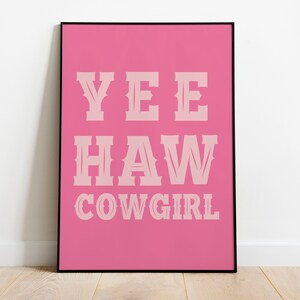 May include: A rectangular wall art print in a thin black frame featuring a vibrant pink background. The text YEE HAW COWGIRL is printed in a light pink, western-style serif font, centered on the print in three distinct lines of bold lettering.