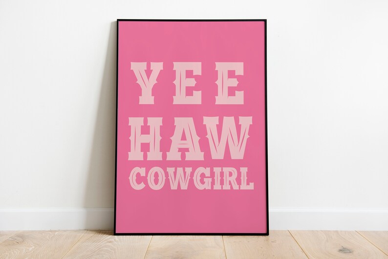 May include: A rectangular wall art print in a thin black frame featuring a vibrant pink background. The text YEE HAW COWGIRL is printed in a light pink, western-style serif font, centered on the print in three distinct lines of bold lettering.