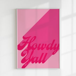May include: A rectangular wall art print in a thin silver frame featuring a two-tone pink background divided diagonally. The text Howdy Yall is printed in a bold, dark pink cursive font with a drop shadow effect across the center of the print.