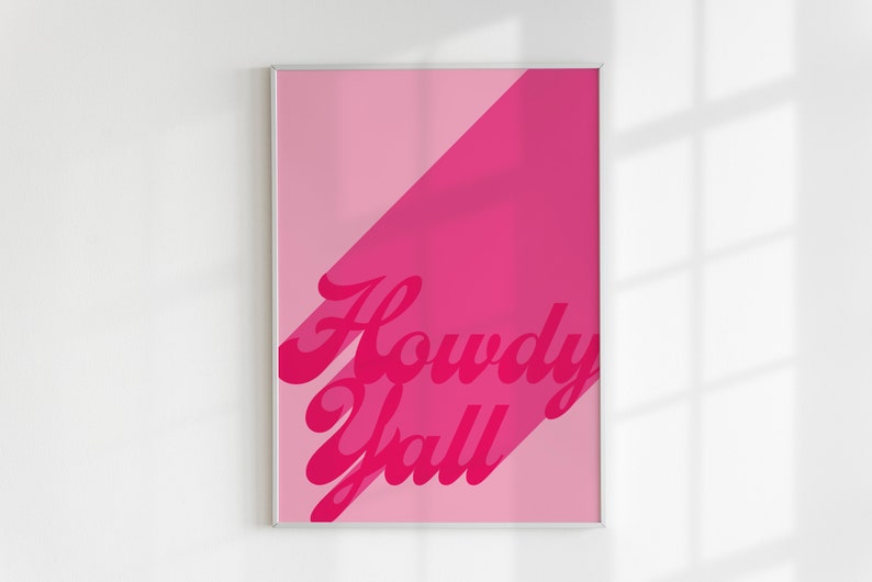 May include: A rectangular wall art print in a thin silver frame featuring a two-tone pink background divided diagonally. The text Howdy Yall is printed in a bold, dark pink cursive font with a drop shadow effect across the center of the print.