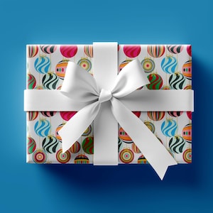 May include: A rectangular gift box wrapped in patterned paper featuring colorful Christmas ornaments. The box is tied with a white ribbon and bow, set against a solid blue background. The ornaments have stripes and swirls.