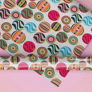 May include: A roll and sheet of patterned wrapping paper featuring colorful, stylized ornaments. The ornaments are in shades of pink, blue, green, orange, and black, with gold string details, on a white background. The paper is set against a pink backdrop.
