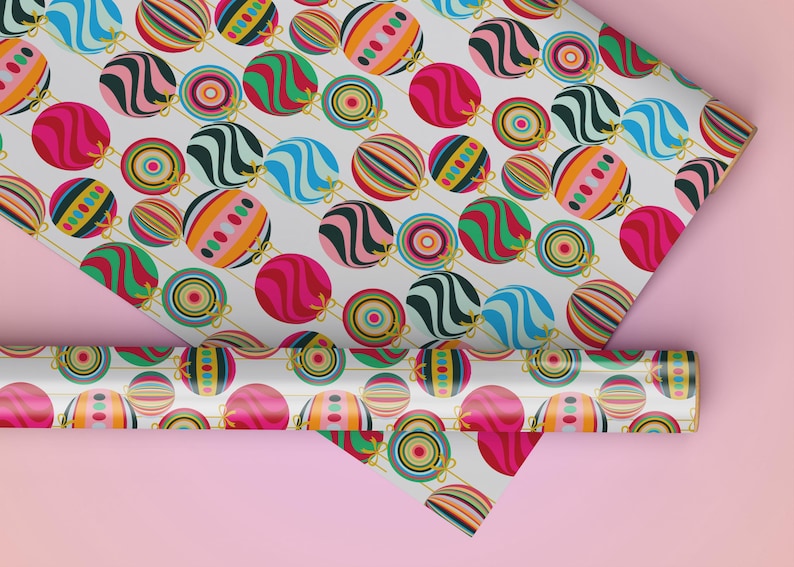 May include: A roll and sheet of patterned wrapping paper featuring colorful, stylized ornaments. The ornaments are in shades of pink, blue, green, orange, and black, with gold string details, on a white background. The paper is set against a pink backdrop.