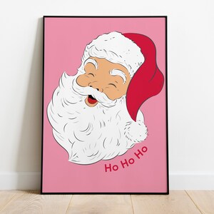 May include: A framed print featuring a cartoon illustration of Santa Claus with a red hat and white beard, set against a pink background. The words "Ho Ho Ho" are written in red. The artwork is displayed on a wooden floor.