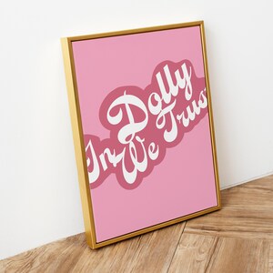May include: A rectangular wall art print in a gold frame featuring a pink background with the white text In Dolly We Trust written in a stylized script font. The text is layered over a darker pink, cloud-like shape on the pink background.