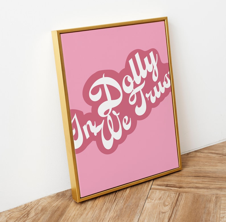 May include: A rectangular wall art print in a gold frame featuring a pink background with the white text In Dolly We Trust written in a stylized script font. The text is layered over a darker pink, cloud-like shape on the pink background.