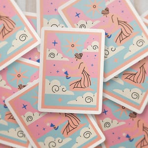 Op de afbeelding: Een verzameling tarotkaarten met een pastelkleurenpalet. Elke kaart toont een hemels ontwerp met een zon, wolken, sterren en een figuur. De kaarten hebben een witte rand en een lichtroze, blauwe en perzikkleurige kleurstelling.