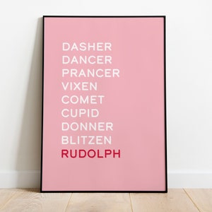 May include: A framed pink art print featuring the names of Santa's reindeer in white and red text. The names are arranged vertically against a solid pink background, with "Rudolph" in red. The frame is black.