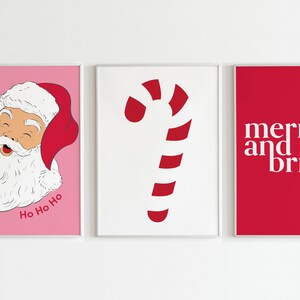 May include: Three framed Christmas-themed prints. The left print features Santa Claus on a pink background with "Ho Ho Ho." The center print shows a red and white candy cane on a white background. The right print has "merry and bright" on a red background.