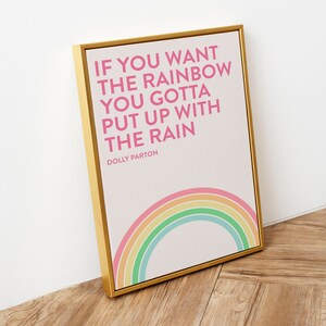 May include: A rectangular wall art print in a gold frame featuring the pink text IF YOU WANT THE RAINBOW YOU GOTTA PUT UP WITH THE RAIN DOLLY PARTON on a white background with a pastel colored rainbow graphic at the bottom of the print.