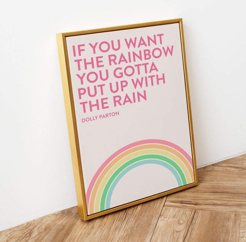 May include: A rectangular wall art print in a gold frame featuring the pink text IF YOU WANT THE RAINBOW YOU GOTTA PUT UP WITH THE RAIN DOLLY PARTON on a white background with a pastel colored rainbow graphic at the bottom of the print.