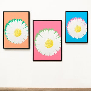 Puede incluir: Tres impresiones artísticas enmarcadas de margaritas. Cada impresión presenta una gran margarita blanca con un centro amarillo y un contorno verde, sobre un fondo vibrante diferente: melocotón, rosa y azul. Los marcos son negros.