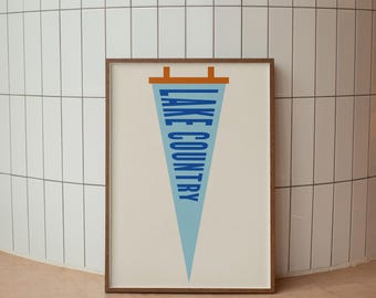 May include: A framed print featuring a light blue triangular pennant with the text LAKE COUNTRY in bold blue capital letters. The pennant has a brown horizontal bar at the top with two small brown tabs extending upward, all set against a cream background.