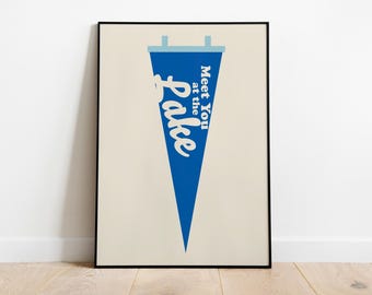 May include: A framed wall art print featuring a blue triangular pennant on a cream background. The pennant displays the white text "Meet You at the Lake" in a stylised font, with light blue accents at the top header and two small hanging tabs.