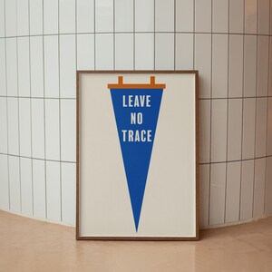 May include: A framed wall art print featuring a blue triangular pennant shape on a cream background. The text LEAVE NO TRACE is printed in white capital letters inside the pennant. The pennant has a brown horizontal bar at the top with two tabs.