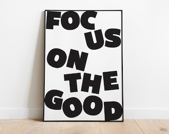 Op de afbeelding: Zwart ingelijste print met de tekst "FOCUS ON THE GOOD" in dikke, zwarte hoofdletters op een witte achtergrond. De letters zijn licht schuin, wat een dynamisch effect creëert. Het kunstwerk staat op een lichte houten vloer.