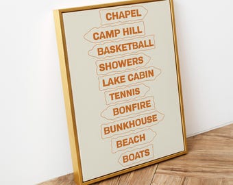May include: A framed vertical wall art print featuring a series of stacked, orange-outlined directional sign shapes on a light cream background. The signs contain the orange text: CHAPEL, CAMP HILL, BASKETBALL, SHOWERS, LAKE CABIN, TENNIS, BONFIRE, BUNKHOUSE, BEACH, BOATS.