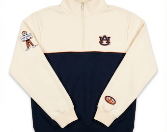 May include: Cream and navy blue quarter-zip sweatshirt. The top half is cream with a navy blue bottom half. Features the Auburn University logo and a cartoon figure on the sleeves. The sweatshirt has a mock neck and a zipper.