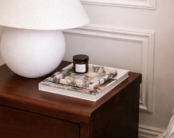 May include: A square marble tray with a grey and white checkered pattern sits atop a stack of books. A small amber glass jar with a black lid and a white label is placed on the tray, alongside a pair of small gold-toned metal hoop earrings.