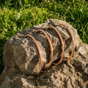 May include: Four copper-coloured, textured bangles rest on a rough, light brown stone. The bangles are curved and appear to be made of a solid metal. The background is blurred, showing green grass and sunlight.