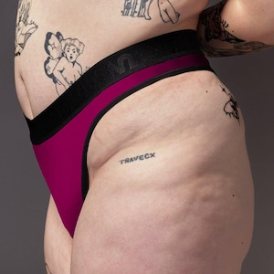 Könnte beinhalten: Nahaufnahme eines magentafarbenen Tangas mit schwarzem Bund. Das Wort "TRAVECX" ist auf die Hüfte tätowiert. Die Haut der Person hat mehrere Tätowierungen. Der obere Teil eines schwarzen Sport-BHs ist sichtbar.