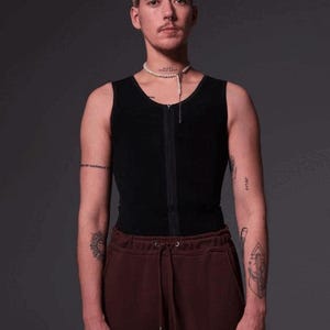 May include: A person wearing a black sleeveless shirt with a zipper and a pearl necklace. They are also wearing maroon sweatpants and have tattoos on both arms.