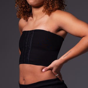 Puede incluir: Un top negro sin tirantes con cierre de cremallera en la parte delantera. El top está hecho de una tela suave y elástica y tiene una silueta ajustada. La modelo lleva pantalones negros y tiene el pelo rizado castaño.