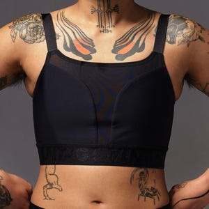 Op de afbeelding: Zwarte sport-bh met een mesh paneel en brede bandjes. De bh heeft een ondersteunend ontwerp en een band met een logo. Het model heeft tatoeages op de borst, schouders en buik. De achtergrond is effen grijs.