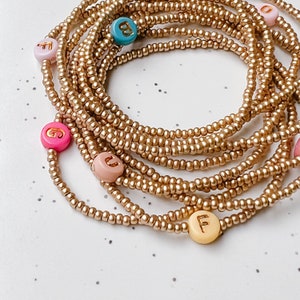Peut inclure: Des perles de taille dorées avec des perles de lettres colorées épelant diverses lettres. Les perles sont disposées en spirale sur une surface blanche. Les perles de lettres sont roses, bleues et jaunes.